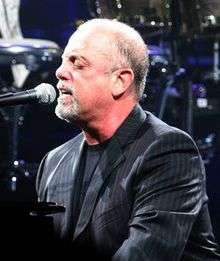 A man with a goatee and his eyes closed behind a microphone on a piano, wearing a black shirt and striped jacket.