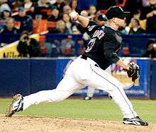 A left-handed man in his mid-thirties wearing a black baseball jersey and cap and white baseball pants throws a baseball from a pitcher's mound.