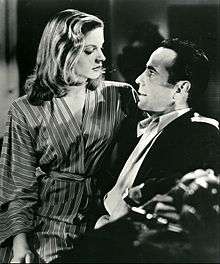 Lauren Bacall in black and white, sitting on Humphrey Bogart's lap facing right in the film To Have and Have Not in 1944