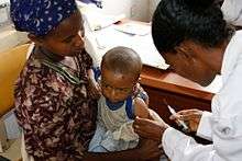 A woman holds a baby while another woman prepares to inject a vaccine.