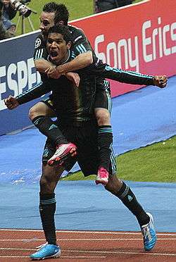 A footballer in a black kit and blue boots celebrates his goal with the cameras on the side of the pitch. A smaller teammate has jumped onto his back.