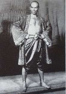 Black and white photo of a man with a shaven head in silky Asian garb; his chest is exposed and his feet are bare; he stands with hands on hips, glowering at the camera