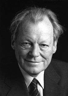 A smiling, older man in a dark suit with a narrow-striped shirt and wide-striped tie. He has receding but long, wavy hair.