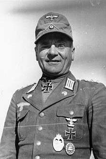A man wearing a field cap and military uniform with various military decorations including an Iron Cross displayed at the front of his uniform collar.