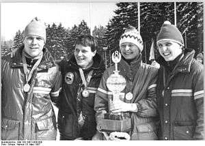 Four smiling people carrying medals around their neck are aligned shoulder-to-shoulder in an outdoor location with pine trees in the background. On the left, a man wears a shiny jacket and winter cap. Next to him, a second man with short dark hair wears a dark jacket with a badge. The third person is a woman wearing a jacket and an embroidered winter cap, and holds a trophy in her hands. The last person, on the right, is another man, also wearing a jacket and a winter cap.