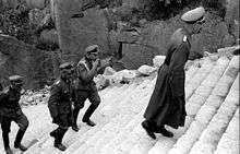 Four men dressed in SS uniforms climb the stone-cut stairs of death, one of them is smoking a cigarette.