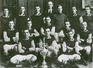 Fourteen footballers pose for a team photograph with a silver trophy in front of them. The team manager stands behind the players.