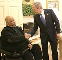 Two men in suits, facing one another and shaking hands; one is seated in a wheelchair.