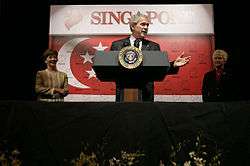 George W. Bush, flanked by a man and a woman, is standing in front of the flag of Singapore, which had been watermarked with numerous small lion symbols.