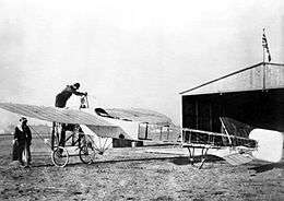 Single-engined monoplane on airfield in front of hangar with Union Jack on flagpole