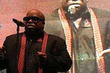 An African-American man wearing sunglasses sings into a microphone. In the background, the image of the same man is projected.