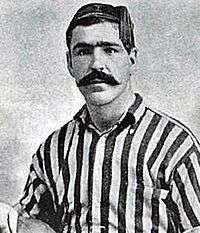 Head and upper torso of a young dark-haired man with thick eyebrows and a large moustache. He is looking straight ahead, and is wearing a tasselled cap and open-necked striped sports shirt.