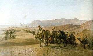 A large number of men mounted on camels in a treeless desert. There is a town in the distance, and bare rocky mountains beyond.
