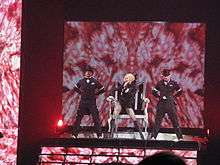 A blond female singing on stage. She wears a black top and long boots on her legs and holds a microphone to her mouth with her left hand. Four male performers in black hats and tail coats flank her. The backdrop display a long array of pink candy in circular motion.