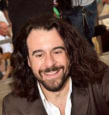head and shoulders view of a smiling man with a dark beard and long hair, in a dark jacket with open white collar