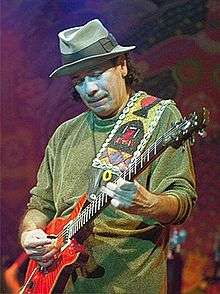 A man wearing a greet hat and shirt, looking down at the guitar he is holding.