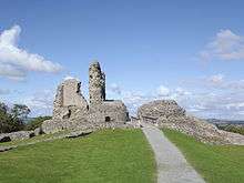 Grey stone ruins, with tall upstanding remains of tower, on a grassy hill, pictured against blue sky.