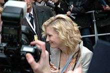 A blonde-haired woman wearing a beige jacket, surrounded by press photographers.