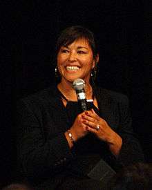 A woman with short black hair smiles as she speaks into a microphone. She is wearing a black blouse and coat.
