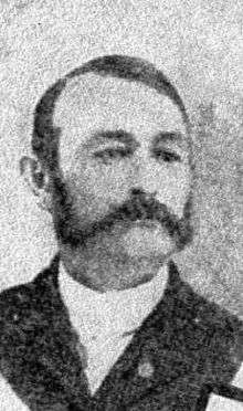 Portrait of a white man with a combover and sideburns connected to a mustache, wearing a dark suit.