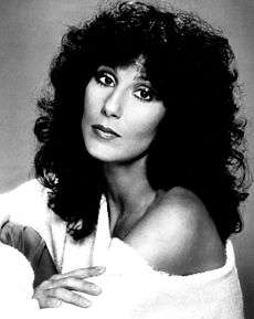 Cher with black curly hair, wearing a white dress