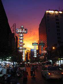 A street during sunset lined with many stalls and shops with a lot of signs bearing Thai and Chinese names