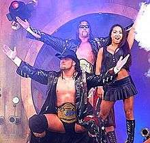An Asian female with dark hair standing next to two male wrestlers wearing wrestling gear, black coats, and championship belts.