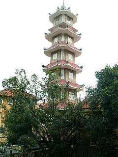 A bell tower that has seven levels of the same pattern, is octagonal with alternating long and short sides, has ornate tiling, and is surrounded by plants.