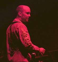 A man with a skinhead haircut sat in front of a musical keyboard. He is looking to his right, away from the camera and is shown side-on. He is wearing a jacket with embroidering on the right sleeve and is holding a drinks can in his right hand. He is bathed in red stage lights.