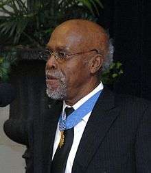 A gray-haired black man standing at a microphone, wearing a medal on a blue ribbon around his neck.