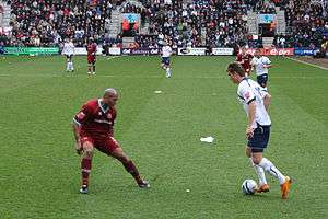 A striker in a white and navy blue soccer strip has the ball. The defender, wearing a claret strip, attempts to make the tackle.