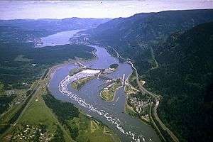 Aerial view of a large river winding through a mountainous gorge. It passes over a dam stretching in four segments from bank to bank across three intervening islands. Highways, passing by clusters of buildings here and there on both banks, run parallel to the river. Whitewater and foam curl downriver from one of the central segments.
