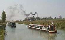 A straight section of canal going into the distance, with two passing  narrow boats in the foreground, with a large brick building and tall chimney in the middle-distance on the right bank of the canal. Smoke is billowing from the chimney, blowing across the canal to the left.