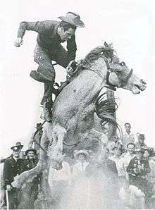 A man dressed in cowboy-style is in mid-air above the back of a horse bucking in a cloud of dust