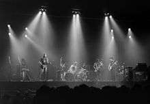 A monochrome image of members of the band. The photograph is taken from a distance, and is bisected horizontally by the forward edge of the stage. Each band member and his equipment is illuminated from above by bright spotlights, also visible. A long-haired man holds a guitar and sings into a microphone on the left of the image. Central, another man is seated behind a large drumkit. Two men on the right of the image hold a saxophone or a bass guitar and appear to be looking in each other's general direction. In the foreground, silhouetted, are the heads of the audience.
