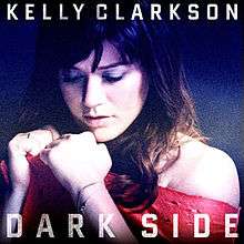 An image of a woman, dressed in pink no-shoulder top, displays her fists in front of a blue background. The words "Kelly Clarkson" (above) and "Dark Side" (below) are written in white semi-transparent, upper-case letters.