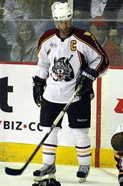 A Caucasian ice hockey player standing on the ice looking down. He wears a white helmet, white jersey with maroon shoulder and arm stripes, black shorts, and white socks. The jersey has a stylized wolf head with a stick and puck behind it as the logo.