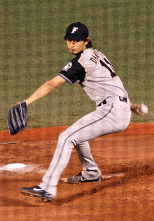 A young Japanese man wearing a grey baseball uniform takes a step forward as he begins to throw a baseball.