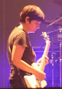 A woman performing onstage with a white bass guitar. She is illuminated by blue lighting.