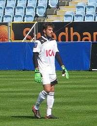 A photograph of a man wearing a white football shirt, green goalkeeper gloves, white shorts and white socks.