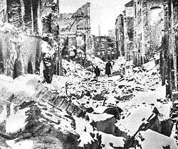 Black and white photo of ruins of buildings in Warsaw. In the distance, two black figures can be seen walking through the rubble.