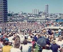 A large crowd has gathered in an open space in an urban setting (tall buildings are visible).  Far away can be seen a platform, and a banner reading SHAME FRASER SHAME.