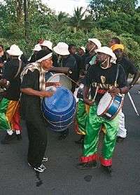 Band members wearing black T-shirts and white hats play drums in the street. Tropical foliage in the background.