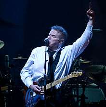 A man wearing a black tie and white dress shirt with an electric guitar hanging from a strap around his neck. His left arm is raised, with his index finger pointed upwards. In the background is a drum set.