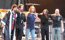 Five male musicians standing on stage. They are standing in an arc, and their hands are on each other's shoulders.