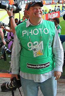 Carey smiling broadly, minus his usual glasses and in casual clothes with a green contest entry top. He is holding a camera with a long lens.