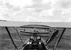 Long front-on view of military biplane on the ground, framed by upper wing, struts, cockpit and twin machine guns of similar machine facing away from camera