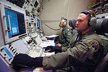 Inside military aircraft. Two personnel wearing green clothes and blue gloves manning communications consoles with wide displays.