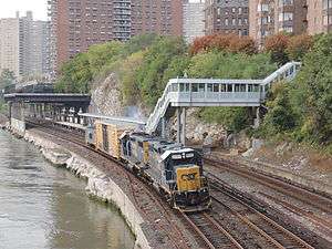 A short freight train, front to the camera, with the letters "CSX" on the front of the locomotive, seen from above next to a station next to water with elevated platforms and a walkway and enclosed stairs connecting it to a nearby street through trees. There are three tracks visible, all with third rails. Behind the station is an area built over the tracks and some brick high-rise apartment buildings