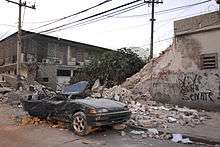 A deep green color car with its roof caved in and the back of the car crushed in. In the background, there is a tree, two phone line poles and a wall with graffiti on it that is half broken down.